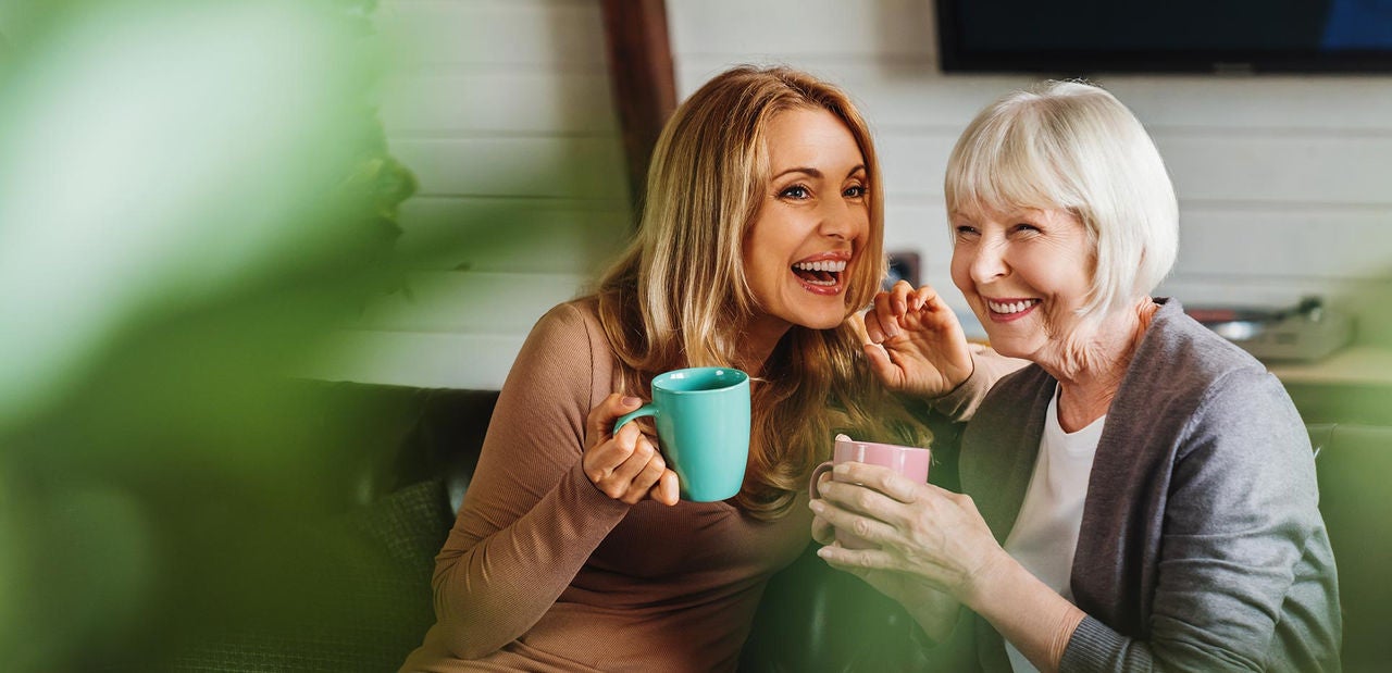 Happy senior mother with adult daughter sitting on couch and holding cups with coffee or tea at home. Togetherness concept