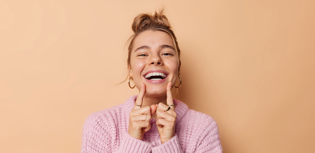 Positive young woman with combed hair points at her broad toothy smile feels very happy has white perfect teeth wears knitted sweater isolated over beige background. People and emotions concept